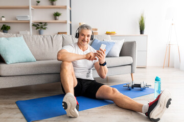 Fototapeta premium Music for workout. Happy middle-aged man with headphones sitting on mat and using smartphone, training at home