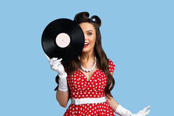 Happy pinup woman in retro dress covering her eye with vinyl record, smiling and looking at camera...