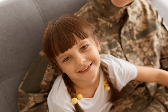Faceless unknown soldier woman wearing camouflage uniform sitting on sofa with charming dark haired female kid with pigtail, kid looking at camera, being glad mother returning from military service.