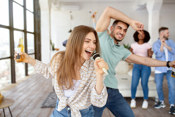 Cheerful Caucasian young lady with bottle of beer singing karaoke on student party with her friends...