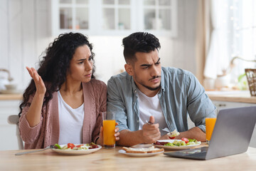 Confused Arab Couple Suffering Problems With Laptop While Eating Breakfast In Kitchen