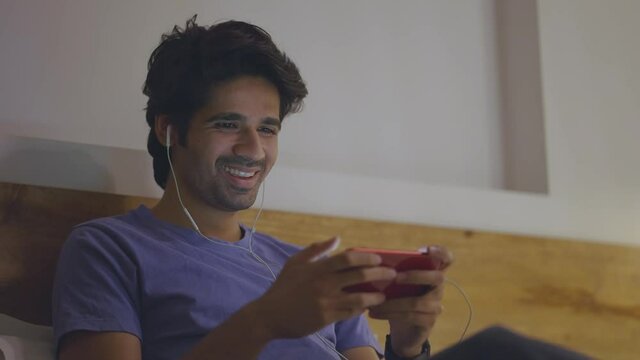 A young happy and cheerful Indian Asian attractive male or man sitting on a bed holding a Mobile Phone in hands using earphones watching a Comedy Movie on Ott platform or Live match sitting at home