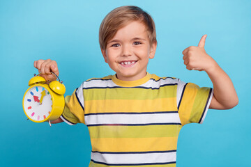 Portrait of attractive trendy cheerful boy holding clock showing thumbup advert isolated over...