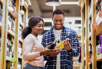 Cheerful black couple holding wine bottles, selecting alcohol beverages at modern supermarket