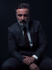 Portrait of adult businessman wearing trendy suit and sitting in modern studio on stylish chair against the black background