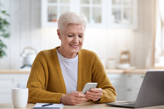 Smiling mature lady sitting at table with laptop, using smartphone