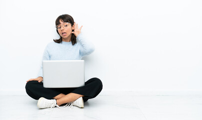 Young mixed race woman sitting on the floor with laptop isolated on white background listening to something by putting hand on the ear