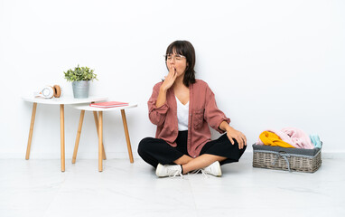 Young mixed race woman folding clothes sitting on the floor isolated on white background yawning and covering wide open mouth with hand