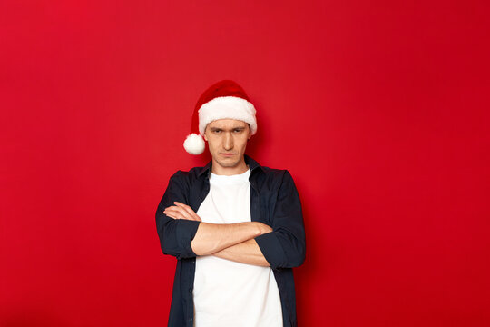 angry disgruntled man folded his arms with cross on chest and offended in Santa Claus Christmas hat. isolated on red background space for text. concept - people, negative emotions, skeptic, hater