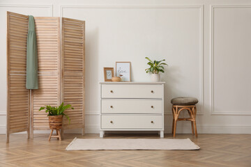 Stylish room with wooden folding screen and chest of drawers near white wall. Interior design