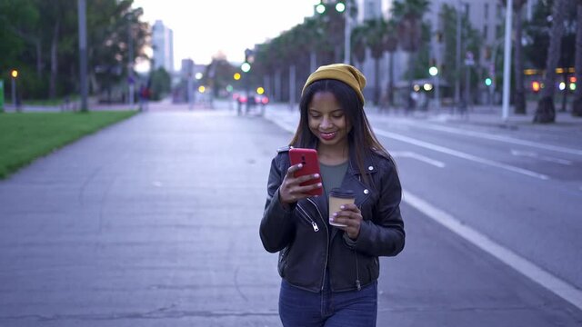Carefree young African American Woman in stylish leather jacket messaging with smartphone in the city in the evening