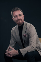 Portrait of adult businessman wearing trendy suit and sitting in modern studio on stylish chair against the black background