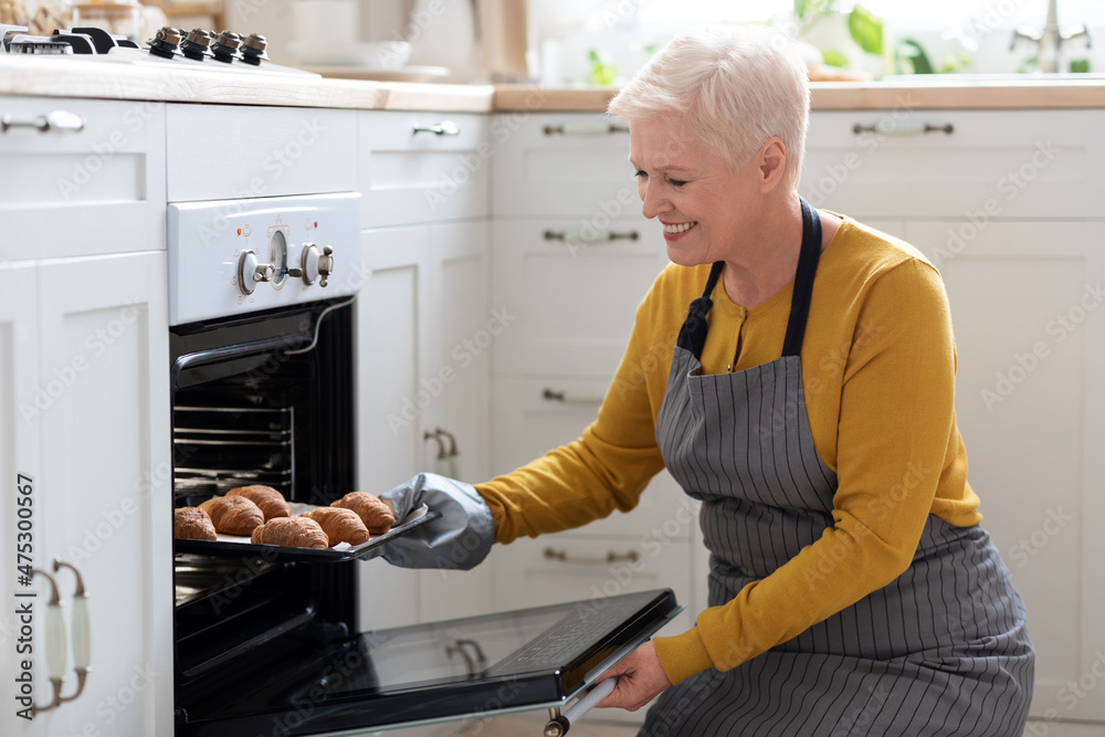 Wall mural smiling elderly woman taking croissants out of oven