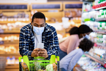 Black guy in face mask checking grocery list on smartphone while his wife and daughter choosing products at mall