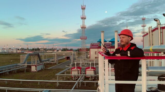 An oil and gas worker, a man with a beard in an orange helmet and overalls, stands on a tower near a rectification tower at an oil refinery. The mechanic speaks on a walkie-talkie