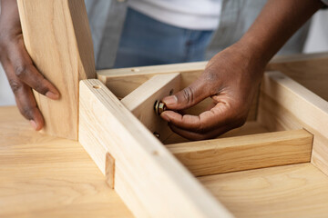 Self-assembly furniture concept. Black man screwing bolt to wooden table, installing desk by...
