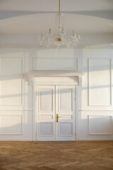 White closed doors in light spacious empty room with classic design and chandelier hanging on...
