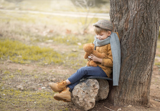 a boy in a yellow jacket, a knitted gray scarf and a vintage tweed cap is sitting by a tree and talking to a teddy bear