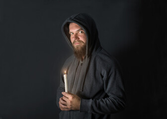 a young man with a beard in black clothes stands in the dark with a burning candle in his hand and looks with a joyful look of madness