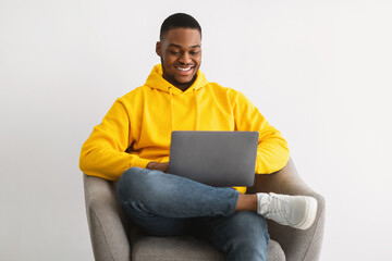 Happy African Guy Using Laptop Sitting In Chair, Gray Background