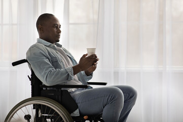 Fototapeta na wymiar Serious adult african american guy disabled in wheelchair holds paper cup with hot drink