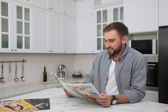 Handsome man reading magazine at white marble table in kitchen