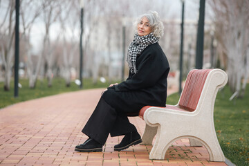 A beautiful mature grey haired lonely woman is sitting on a bench in a cold autumn park