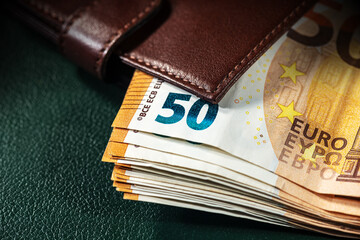 Extreme closeup of a brown leather wallet with a group of European Union banknotes (fifty Euro) on a green background.