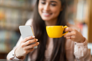 Coffee break. Happy arab lady using smartphone and drinking hot drink in cafe, closeup, selective focus