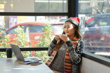 Asian woman relaxing with sip a drink while working with laptop at coffee shop in the winter, concept offsite work or ect