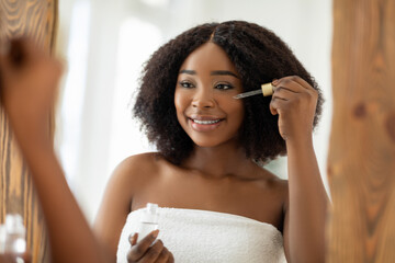 Beauty cosmetics for young skin. Sensual black woman applying regenerating serum onto her face near mirror indoors