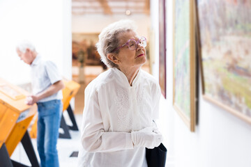 mature woman examines paintings in an exhibition in hall of an art museum