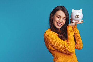Excited pretty girl in knitted orange sweater holding white piggy bank with lots of money near face...