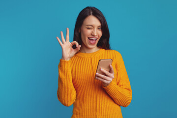Beautiful caucasian woman dressed in warm knitted orange sweater showing ok gesture and blinks eye while using smartphone against blue background