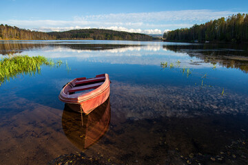 Obraz premium A red old wooden boat anchored near the shore, in a secluded harbor. Landscape photography of a beautiful blue lake and an anchored red boat.