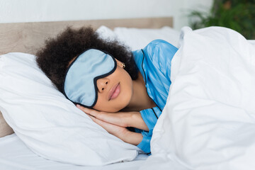 curly african american woman in blue pajamas and silk eye mask sleeping in bed