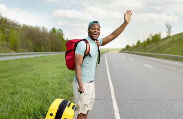Attractive black guy with luggage waving down a car, hitchhiking on roadside, traveling by autostop, outdoors