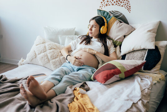Cute young pregnant woman relaxes lying on the bed and listening to music on wireless headphones.