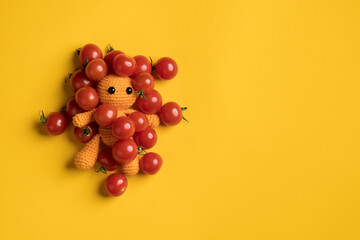 Cute homemade knitted toy man in red cherry tomatoes, on a yellow background. Copy space. Top view. 