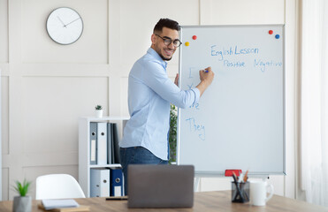 Fototapeta premium Arab male teacher giving online English lesson, writing grammar rules on blackboard. Distance education during covid