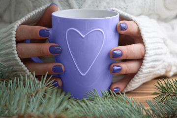 Woman hands in white sweater with purple nail polish hold mug with heart near Christmas tree. Color Year 2022