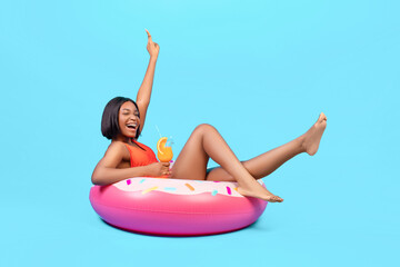 Cool black lady sitting on inflatable ring, having fun, enjoying summer cocktail on blue background, full length