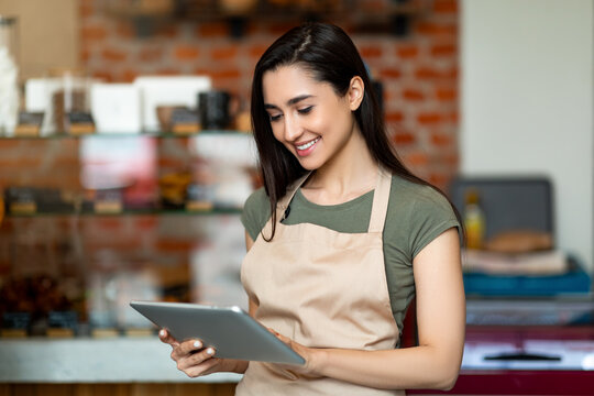 Young female owner of modern cafe in apron holding digital tablet and standing with shopwindow on background