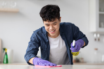 Cheerful asian guy cleaning kitchen table with spray and cloth