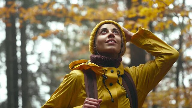 close up portrait of pretty inspired young woman hiker with backpack, wearing bright yellow raincoat and beanie. girl looks around enjoying nature beauty in wonderful autumn woods.