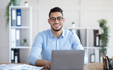 Happy young Arab businessman in glasses sitting at desk, working on laptop at company office