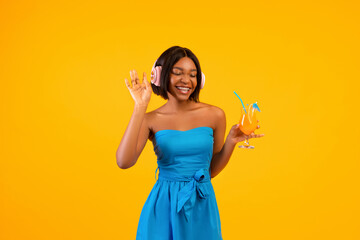 Black lady in stylish outfit holding exotic cocktail and enjoying favorite music in headphones on orange background