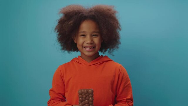 Happy black girl eating chocolate bar holding in hands smiling at camera standing on blue background. Cute kid enjoying milky chocolate indulgence. 