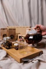 A hand pouring tea from a glass teapot onto a wooden serving tray. A jar of tea and a candle on a textile background.