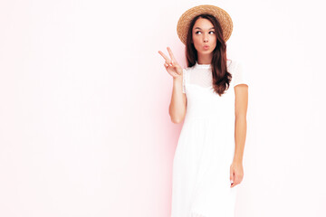 Young beautiful smiling female in trendy summer  clothes. carefree woman posing near pink wall in studio. Positive brunette model having fun.Cheerful and happy.In hat.  Shows peace sign